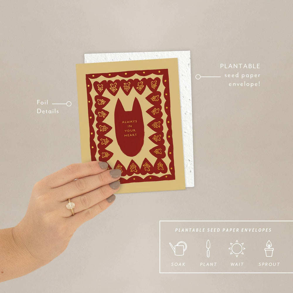 Hand holding a plantable seed paper envelope with red and gold design on a beige background