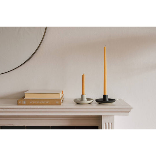 Two candle holders with candles on a white surface, with a mirror and books in the background.