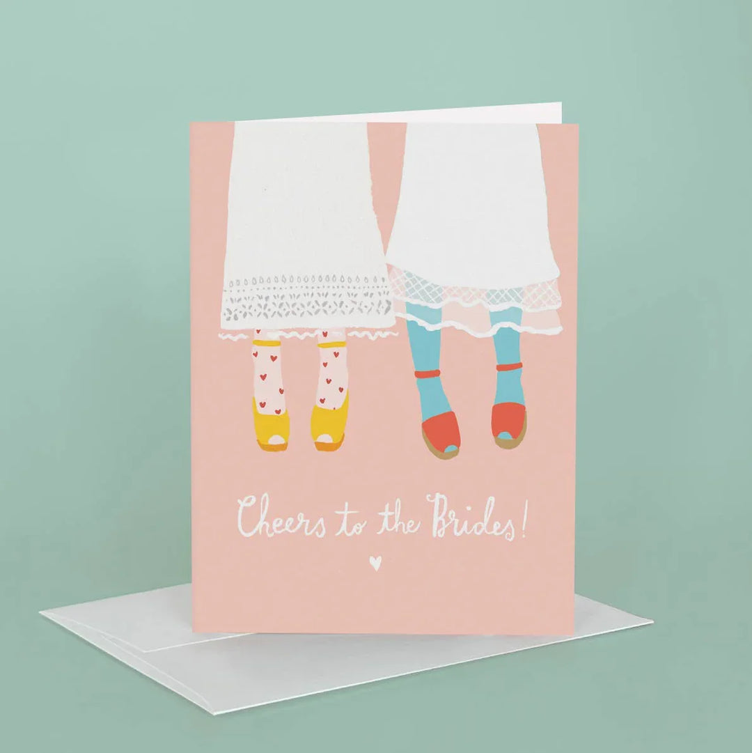 Greeting card with illustration of two women in dresses and shoes, text 'Cheers to the Brides!', on a light pink background.