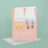 Greeting card with illustration of two women in dresses and shoes, text 'Cheers to the Brides!', on a light pink background.