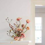 Floral arrangement in a white vase on a white surface with a light gray wall and window in the background.