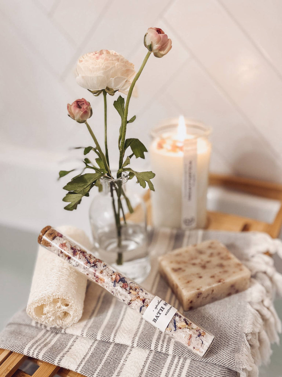 Candle, flowers in a vase, soap bar, and tube of bath salts on a wooden surface with a towel.