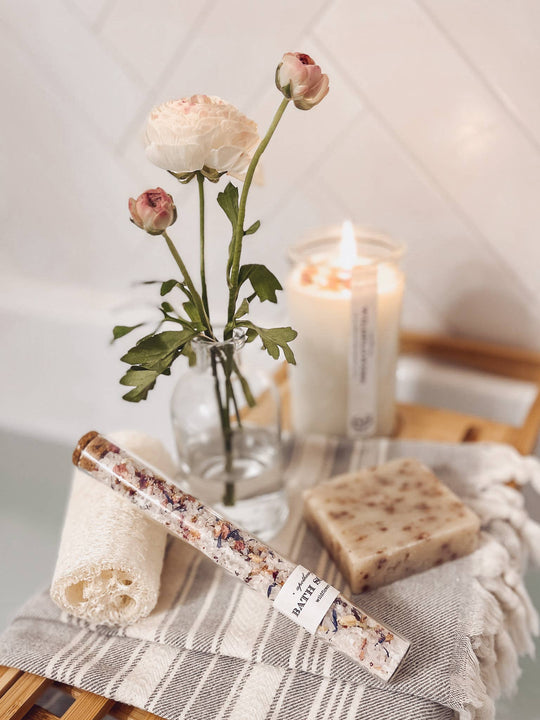 Candle, flowers in a vase, soap bar, and tube of bath salts on a wooden surface with a towel.